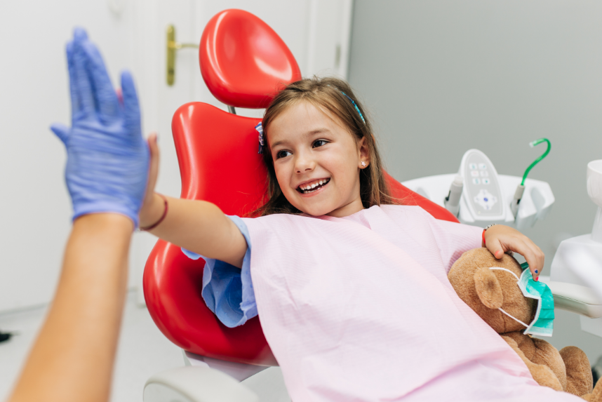 Little girl high fiving dentist with teddy bear wearing surgical mask in dental chair